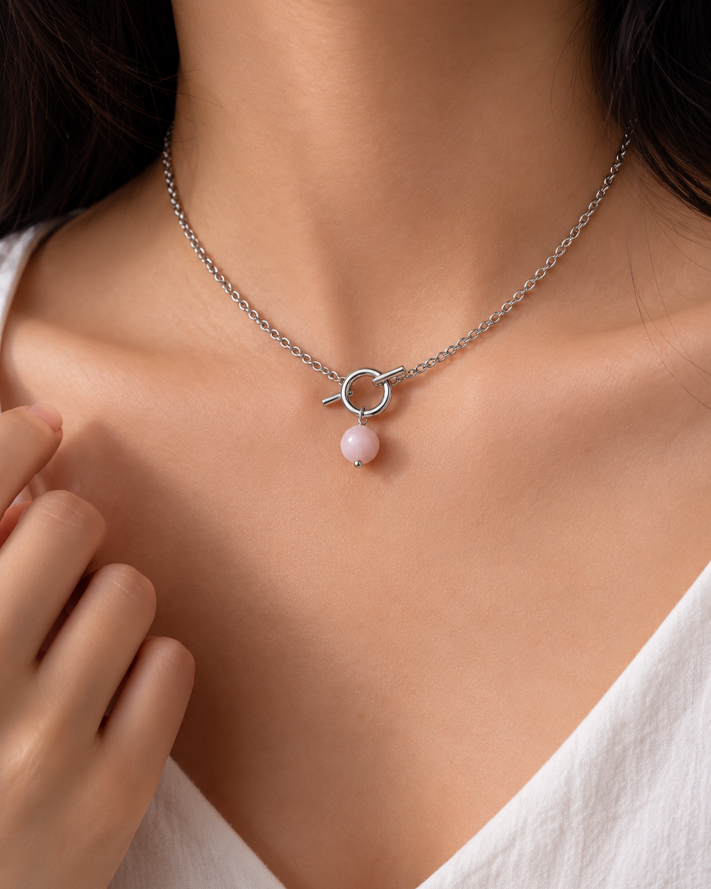 A close-up of a model wearing a delicate silver chain necklace with a small circular ring pendant and a single light pink bead drop.