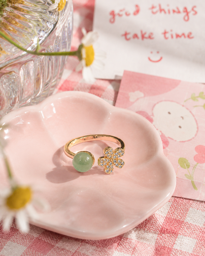 A gold ring with a round light green stone and a flower-shaped crystal cluster setting, displayed on a pink ceramic dish over a pink gingham cloth with daisies and a card in the background.