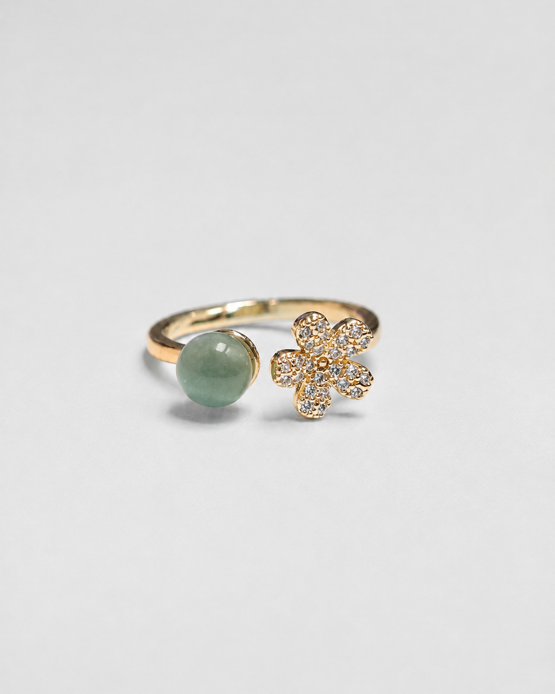 A gold-plated open ring featuring a smooth oval light green stone on one side and a small gold flower cluster paved with clear stones on the other, set against a plain light gray background.