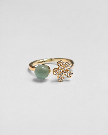 A gold-plated open ring featuring a smooth oval light green stone on one side and a small gold flower cluster paved with clear stones on the other, set against a plain light gray background.